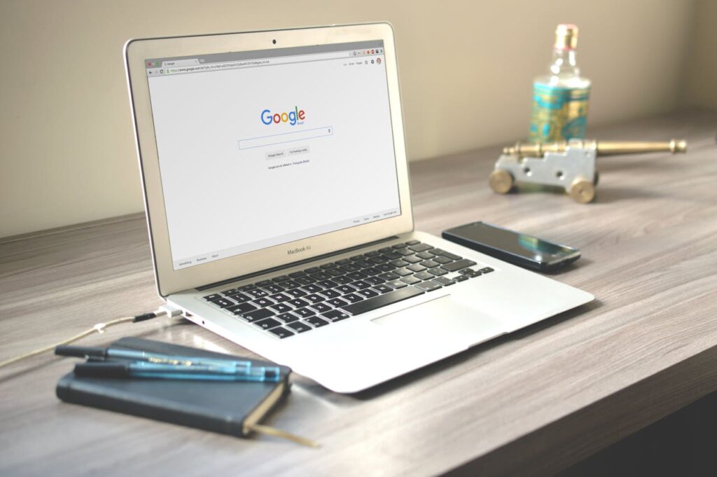 SEO Agency. A neat workspace featuring a laptop displaying Google search, a smartphone, and a notebook on a wooden desk.